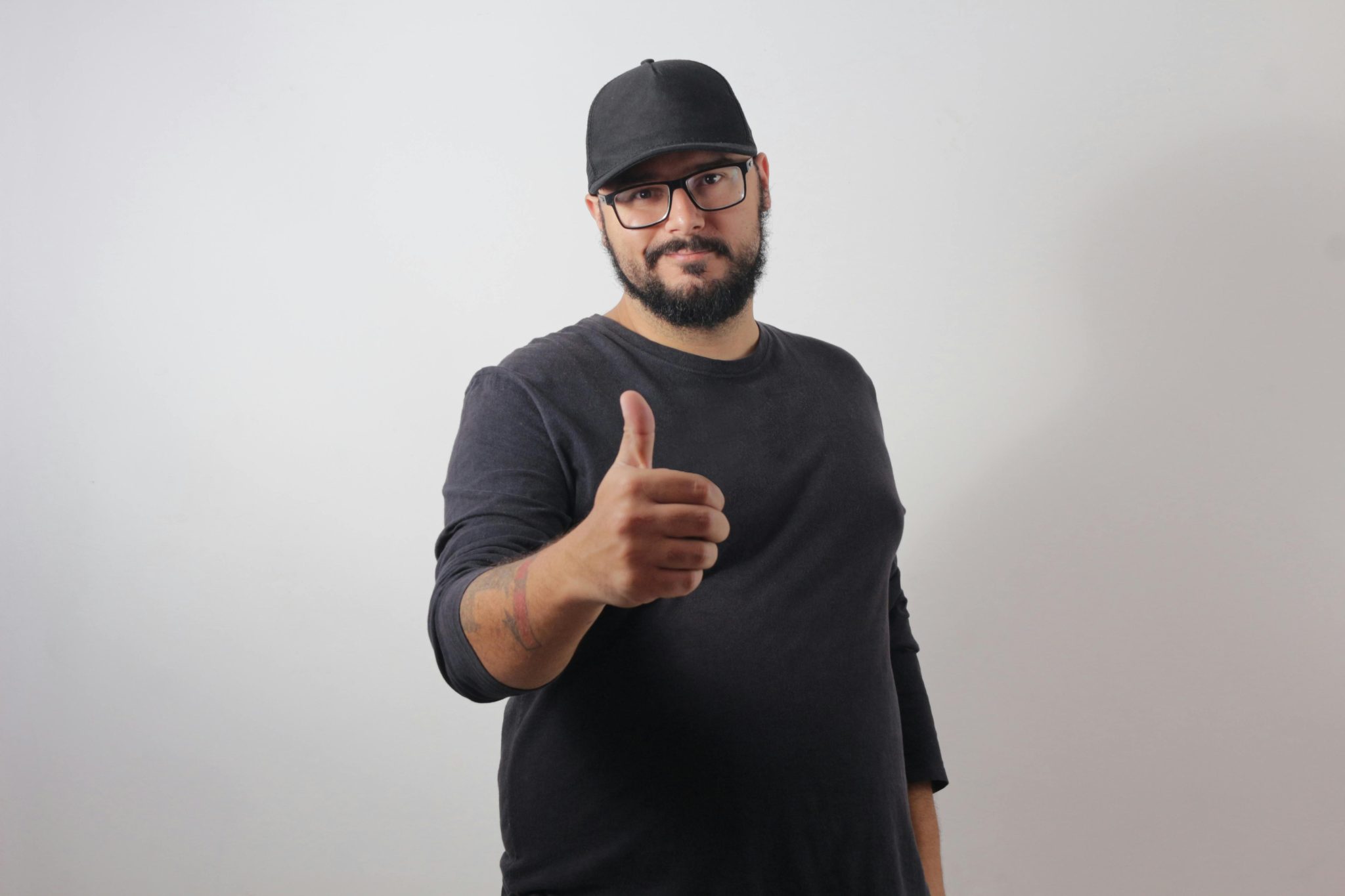 A bearded man in a black shirt and cap gives a thumbs-up gesture with a smile against a white background.